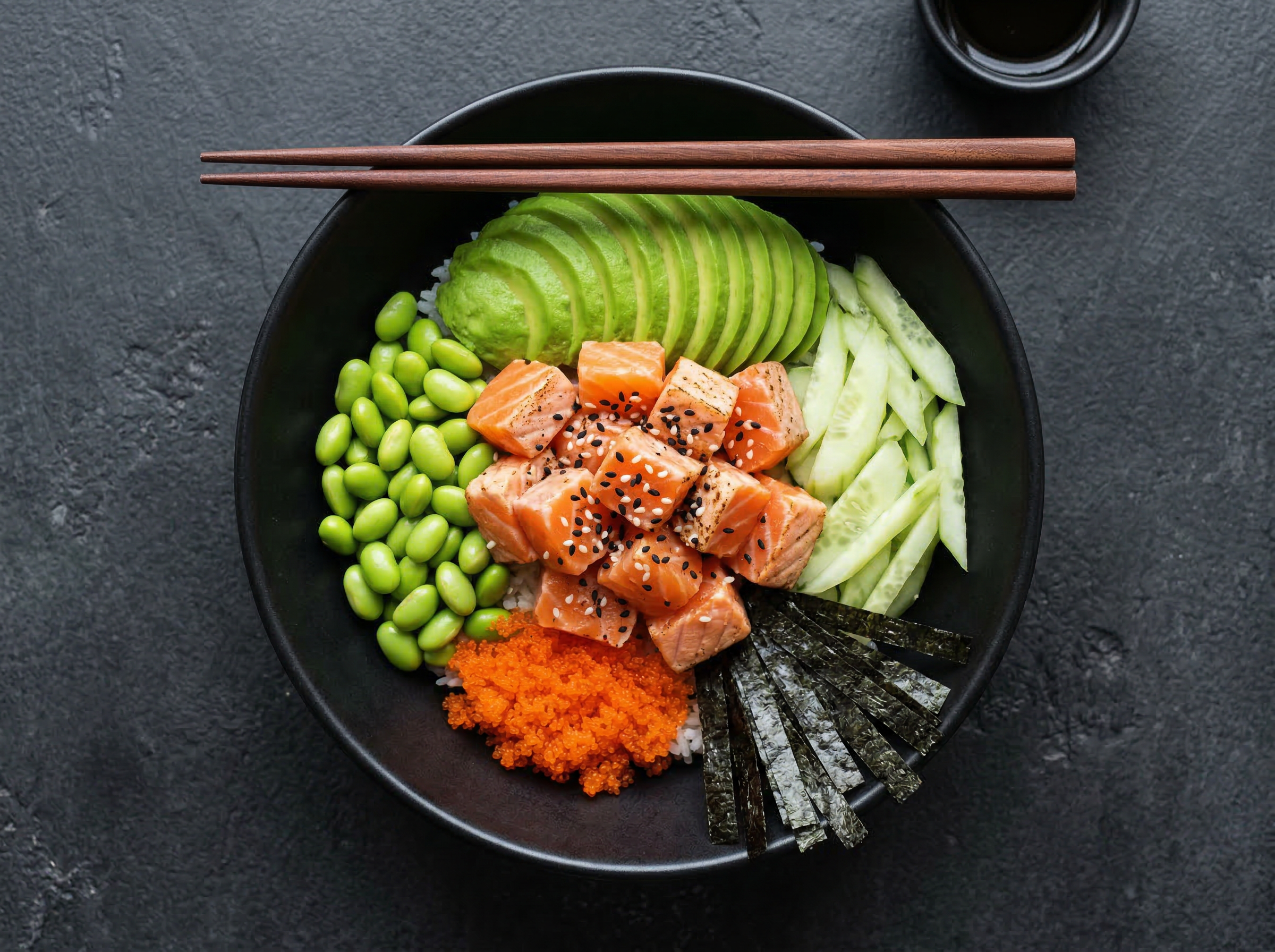 Matte black poke bowl overhead with salmon, cucumber, avocado, dark nori strips, and sesame seeds in clean distinct zones against a high-contrast dark field