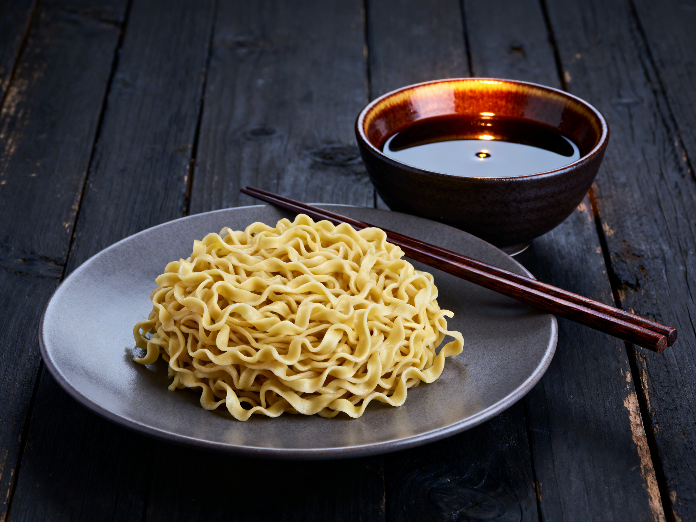 Tsukemen dipping noodles — controlled noodle nest on a flat ceramic plate in the foreground, rich dark dipping broth bowl behind and to the right, dark weathered wood surface, 40-degree elevation