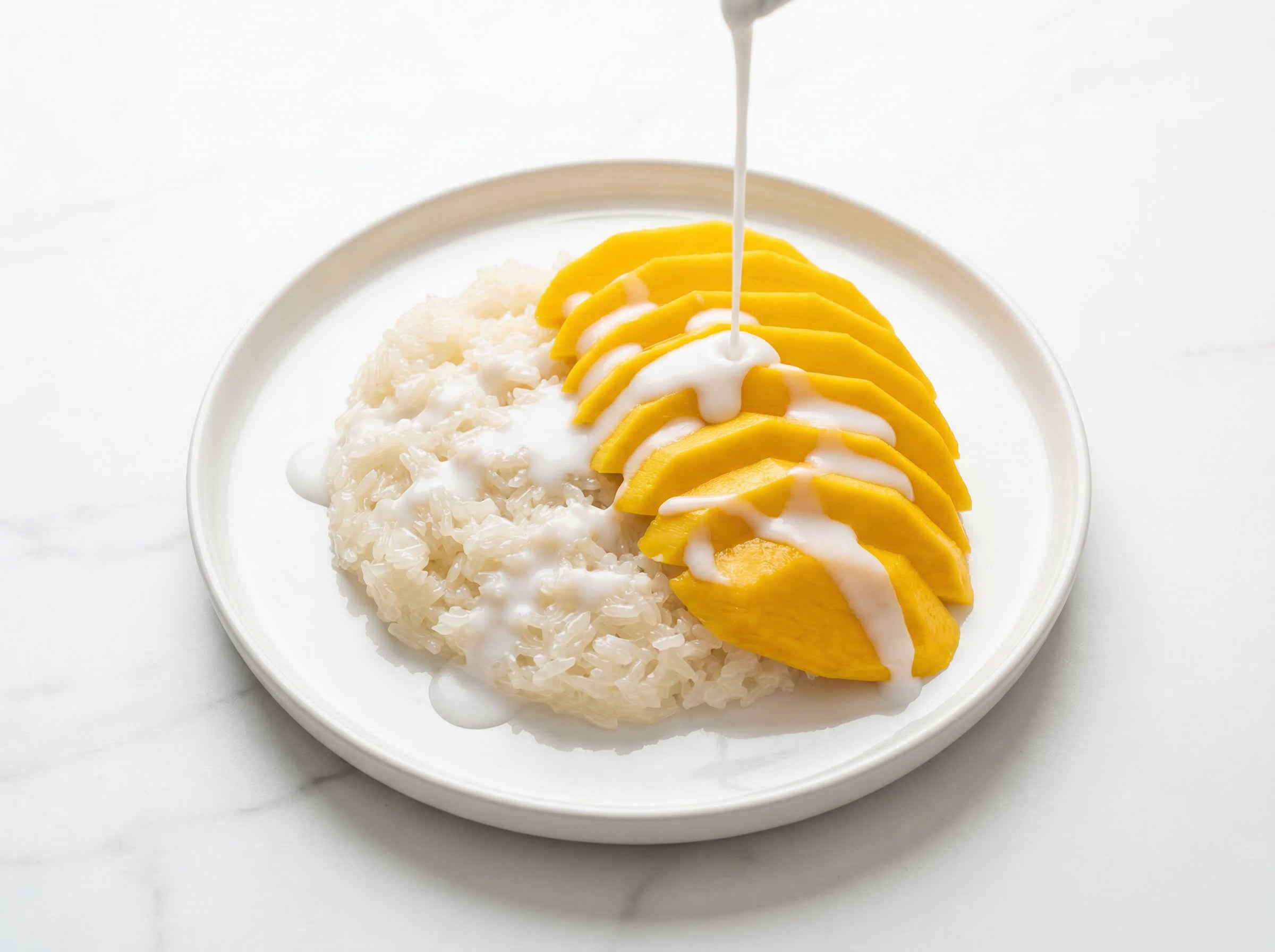 Mango sticky rice photographed from directly overhead, fan-sliced yellow Ataulfo mango arranged on white glutinous rice with coconut cream freshly drizzled in a thin stream still visible mid-pour, clean white surface